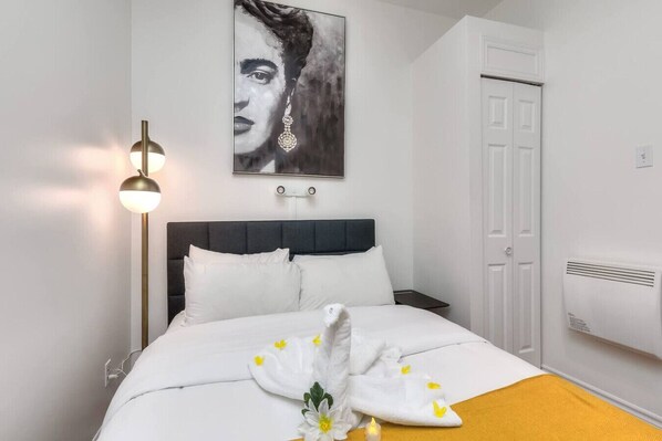 A neatly made bed with white linens, a bedside table, a wall-mounted artwork, a floor lamp, and a white door.