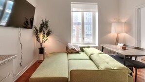 Ein modernes Wohnzimmer mit einer Couch, einem an der Wand montierten Fernseher, einem Fenster mit Blick auf Gebäude und einer Lampe auf einem Tisch.