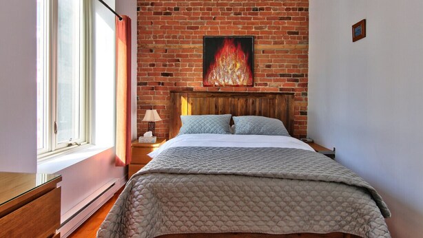 Ein Schlafzimmer mit Backsteinwand, einem Bett mit Steppdecke, einem Nachttisch, einer Lampe und einem Fenster mit Vorhängen.