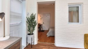A hallway with a white brick wall, a potted plant, a door leading to a room with a bed and a window, and a lamp on the left.