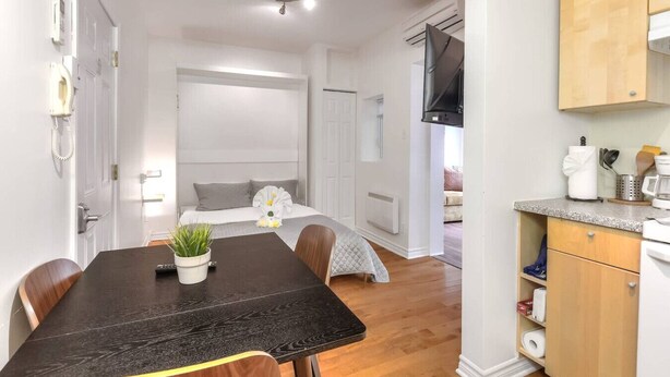A modern hotel room with a dining area, a bed with a gray headboard, a kitchen area, and a wooden cabinet.