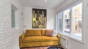 A modern living room with a mustard-colored sofa, a black abstract painting, and a view of a building through a large window.