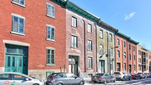 A row of multi-story brick buildings with green-framed windows and doors, parked cars along the street, and a clear blue sky.