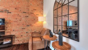 Una habitación con pared de ladrillo, un sillón de cuero, una pequeña mesa de madera con una lámpara y un espejo grande que refleja la pared de ladrillo.
