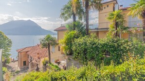 A coastal building with a balcony overlooking the sea, surrounded by lush greenery and a mountain in the background.