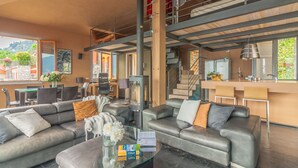 A modern living room with leather sofas, a glass coffee table, and a fireplace. There is a kitchen area with a bar and a view of the outdoors.