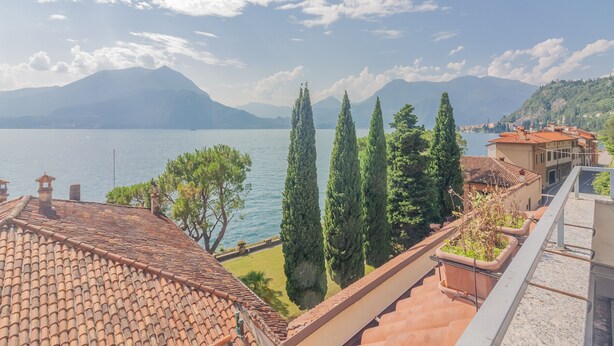Una terraza con vistas a un lago, montañas y altos cipreses.