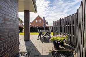 Eine Terrasse im Freien mit einer Backsteinmauer, einem Tisch und Stühlen sowie einem Blumentopf mit violetten Blüten.