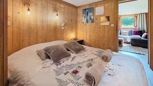A wooden-paneled bedroom with a large bed, a nightstand, a painting, and a view through an open door to a balcony. - The cosy refuge (Chamonix-Mont-Blanc)