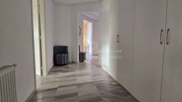 A hallway with a tiled floor, a black chair, and a trash bin.