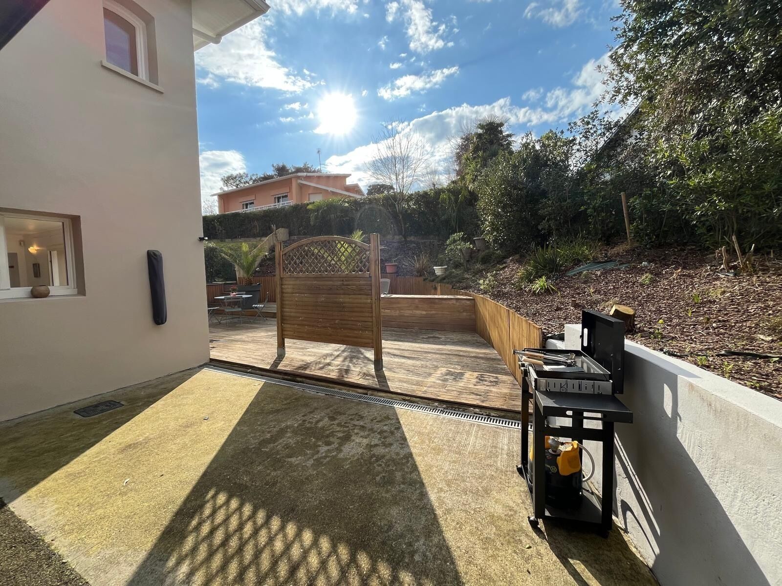 Une terrasse avec une plateforme en bois, un barbecue en métal et une vue sur un jardin et des bâtiments en arrière-plan.