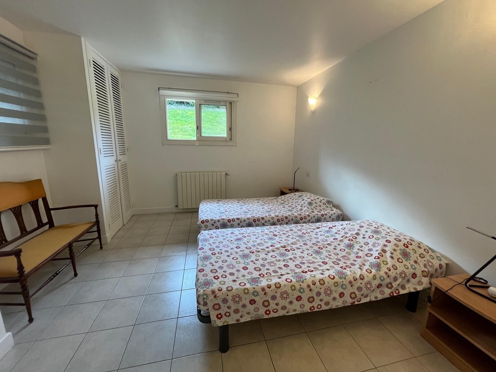 A small, well-lit room with two single beds, a wooden bench, a radiator, and a window with a view of greenery.