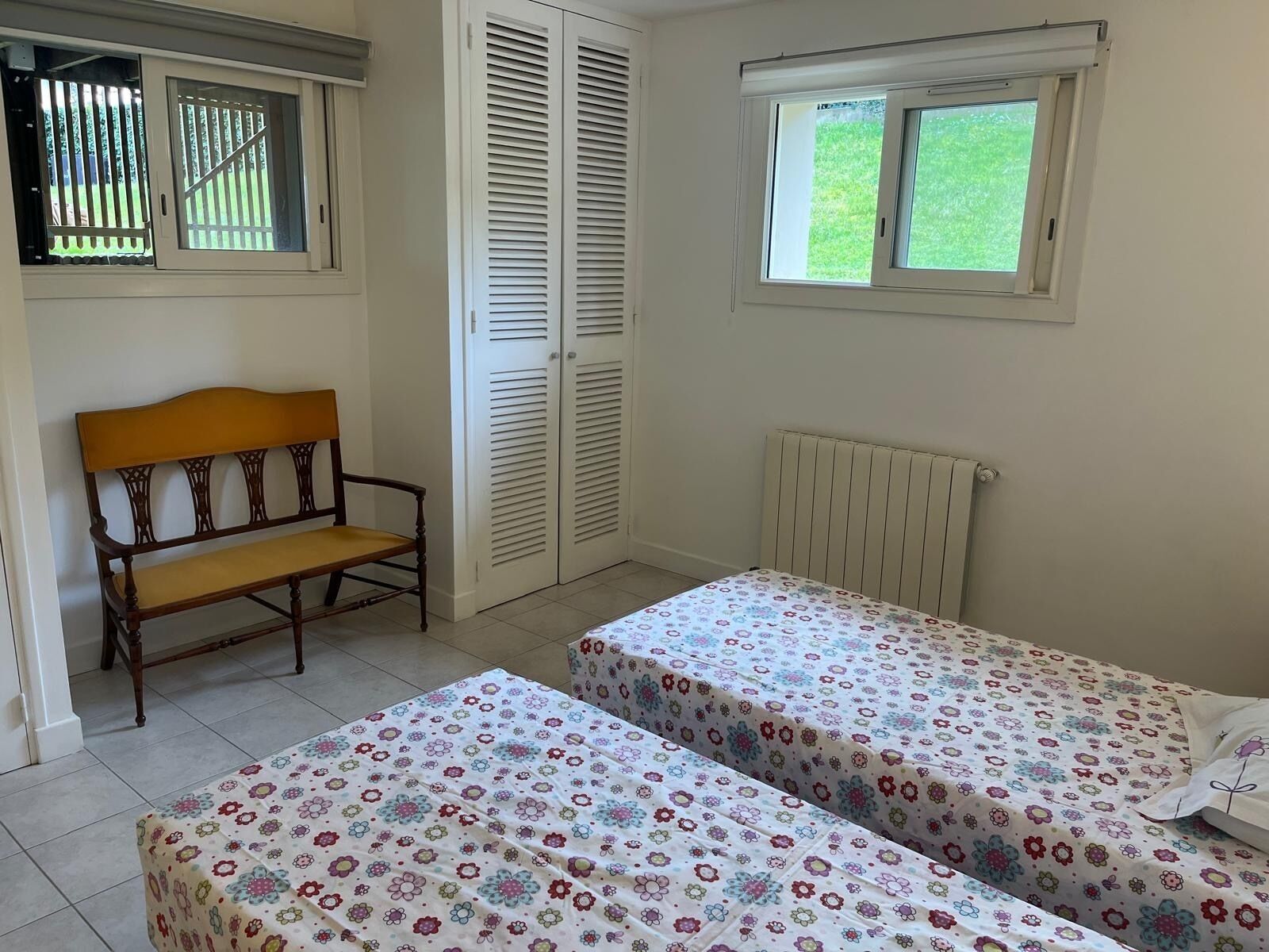Une chambre avec deux lits simples, un banc en bois, un radiateur et une fenêtre avec des stores.