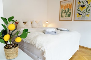 A neatly made bed with a white blanket, a potted plant with yellow flowers, and two framed pictures on the wall.