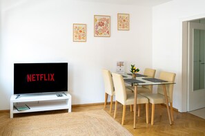 A living room with a flat-screen TV displaying 'NETFLIX,' a glass-top dining table with four chairs, and three framed floral pictures on the wall.