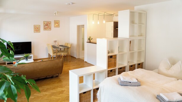 A modern hotel room with a bed, a TV, a sofa, a dining area, and a white shelving unit with baskets. - Cozy apartment+kitchen+Netflix (Essen)