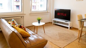 A living room with a leather sofa, a small table with a plant, a TV on a stand, and a dining area with chairs. - Cozy apartment+kitchen+Netflix (Essen)