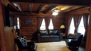 A log cabin interior with a wooden ceiling, a black leather sofa, two armchairs, a wooden table, and a television mounted on the wall. - Authentic Log Cabin in the Southern Ozark Mountains with hot tub (Mountain Home)