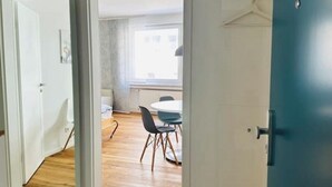 Une porte ouverte donne sur une pièce où se trouvent une table à manger et des chaises, une fenêtre avec des rideaux et une porte bleue.