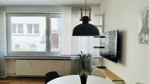 Un espace repas moderne avec une table ronde, des chaises, une plante en pot, un luminaire suspendu noir et un téléviseur fixé au mur.