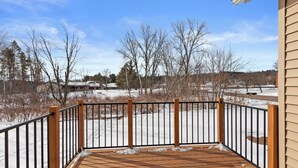 Eine Holzdar Terrasse mit Metallgeländer, mit Blick auf eine verschneite Landschaft mit kahlen Bäumen und Gebäuden in der Ferne.