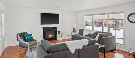 A modern living room with a fireplace, grey sofas, a TV, and large windows.