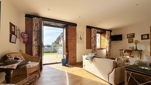 A spacious living room with a wooden floor, a white sofa, a wicker chair, and a dining table. There is a large window with curtains, a fire extinguisher, and a television on the wall. - 🏡 Perfect Island Getaway - Townhouse with Private Parking 🚗✨. (Isla)