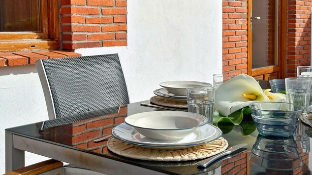 Un área de comedor al aire libre con una mesa para dos, con cubierta de vidrio, sillas de metal y un centro de mesa con flores blancas.