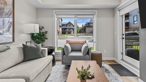 Una sala de estar moderna con un sofá gris, una mesa de centro de madera y vistas a una zona residencial a través de amplios ventanales.