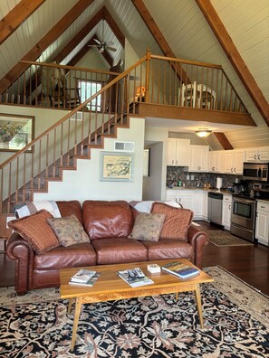 A spacious living room with a brown leather sofa, a wooden coffee table, and a large patterned rug. A staircase with wooden railings leads to an upper level.