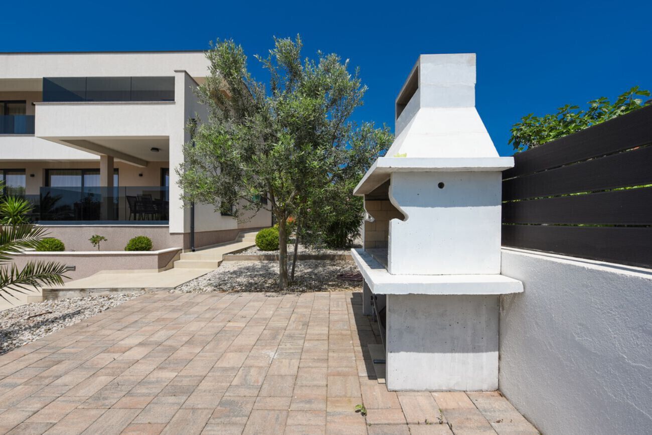 Un espace extérieur moderne, aménagé en patio, avec un barbecue, un petit arbre et un chemin pavé.