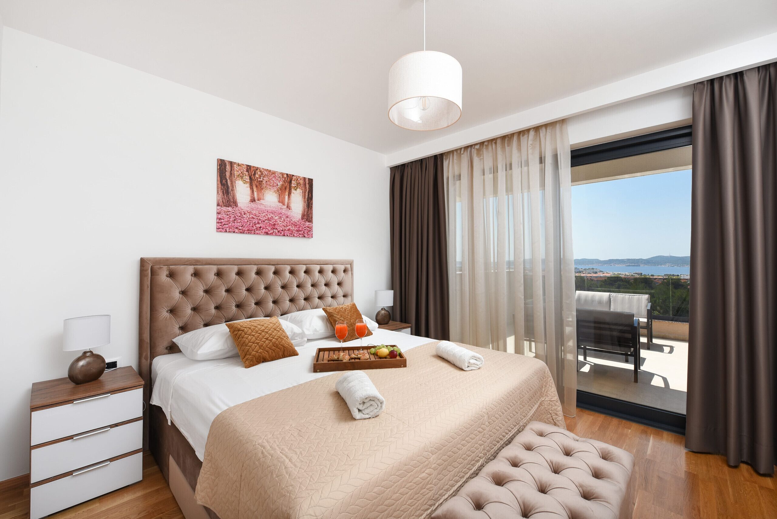 A modern hotel room with a large bed, a nightstand, a tray with fruit, and a view of the outdoors.