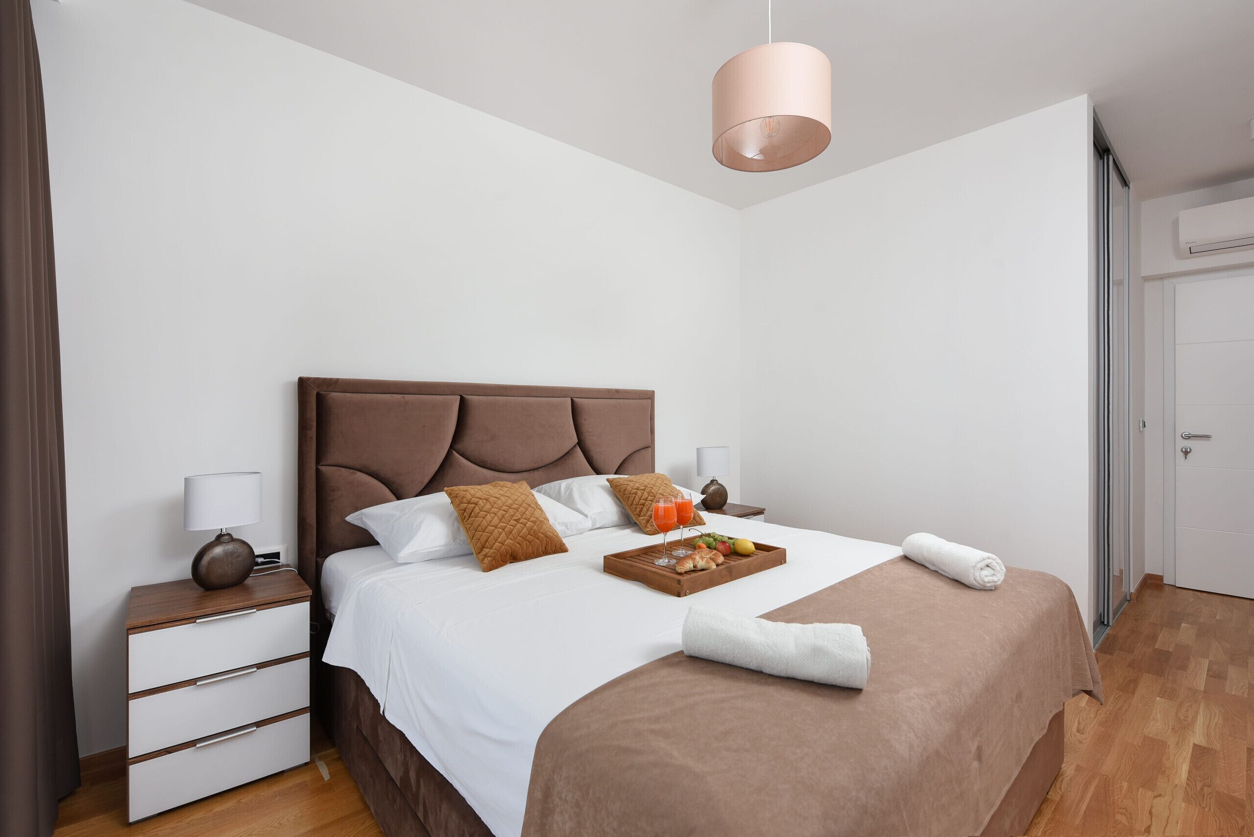 Ein ordentlich bezogenes Bett mit braunem Kopfteil, ein Nachttisch mit Lampe und ein Tablett mit Frühstücksangeboten.