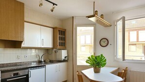 Ett kök med vita skåp, en inbyggd ugn, en diskmaskin, ett matbord med stolar, en krukväxt och ett fönster med utsikt över byggnader.
