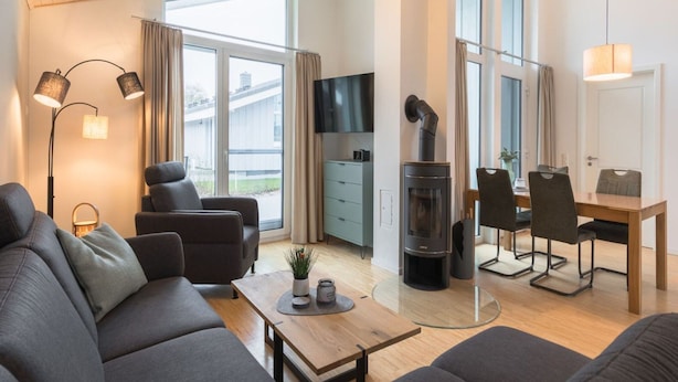 A modern living room with a sofa, a coffee table, a fireplace, and a dining area with chairs and a table. - Ferienhaus mit Sauna im Ferienpark Mirow (Mirow)
