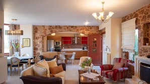 Un salón con un sofá, sillones y una mesa de centro. Se observa una cocina al fondo con armarios rojos y una pared de piedra.