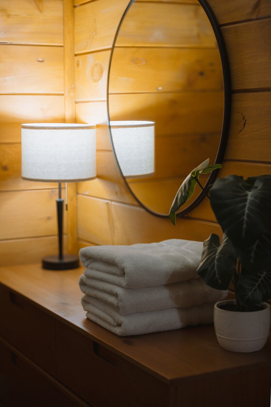 A wooden cabin interior with a mirror, a lamp, a stack of towels, and a potted plant.