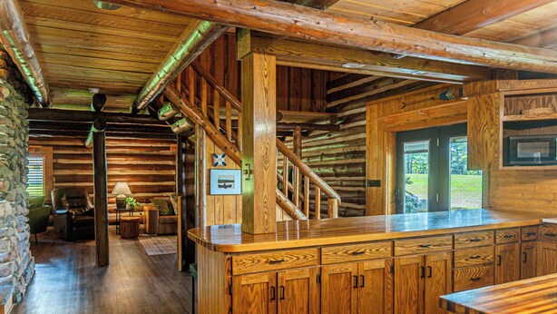 A log cabin interior with a wooden staircase, a kitchen area with wooden cabinets, and a stone wall. - The Little Place on Pine (Harrison)