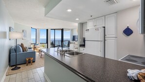 A modern kitchen with a granite countertop, white cabinets, and a built-in oven. There is a dining area with a table and chairs, and a large window offering a view of the ocean. - Sunbird 1BR/1BA, Direct Beachfront! Stunning Views! (Panama City Beach)