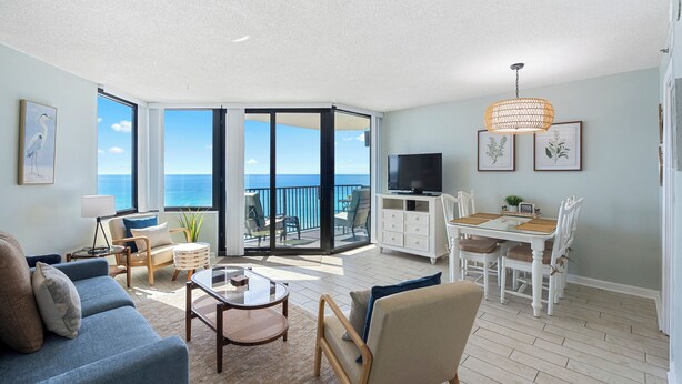 A spacious living room with a large sliding glass door leading to a balcony with a view of the ocean. There is a dining area with a table and chairs, a television, and framed botanical prints on the wall. - Sunbird 1BR/1BA, Direct Beachfront! Stunning Views! (Panama City Beach)