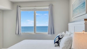A bedroom with a large window offering a view of the sea, a bed with white bedding, and a lamp on a bedside table. - Direct Beachfront, wrap balcony, sleeps 10! Shores of Panama (Panama City Beach)