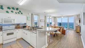 A modern kitchen with white cabinets, a microwave, and a stove. There is a dining area with a round table and chairs, and a balcony with a view of the ocean. - Direct Beachfront, wrap balcony, sleeps 10! Shores of Panama (Panama City Beach)