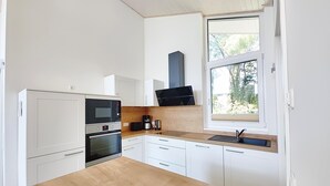 Una cocina moderna con armarios blancos, encimera de madera, horno empotrado, microondas, cafetera y ventana con vistas a árboles.