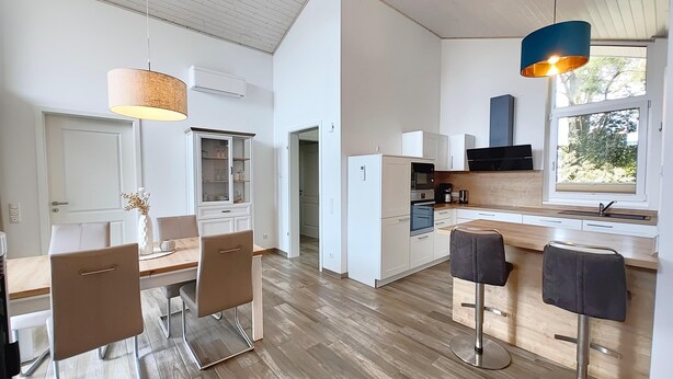 Una cocina moderna con armarios blancos, encimera de madera y horno integrado. Hay dos taburetes de bar y una mesa de comedor con sillas.