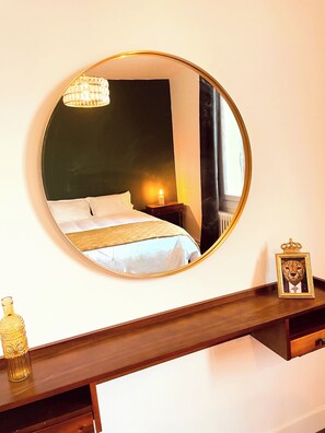 Ein runder Spiegel spiegelt ein Bett mit weißer Bettwäsche, einen Nachttisch mit einer Lampe und einen Bilderrahmen auf einem Holzregal.