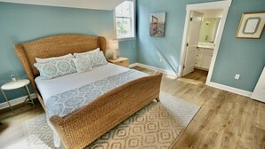 Une chambre à coucher avec un grand lit, une table de chevet, une commode, une fenêtre et une porte donnant sur une salle de bain.
