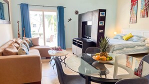 Ett modernt hotellrum med en soffa, ett glasbord, en säng, en TV och en balkong med blå gardiner.