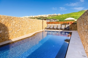 Una piscina con paredes de piedra, cerca de madera y vista a edificios al fondo.