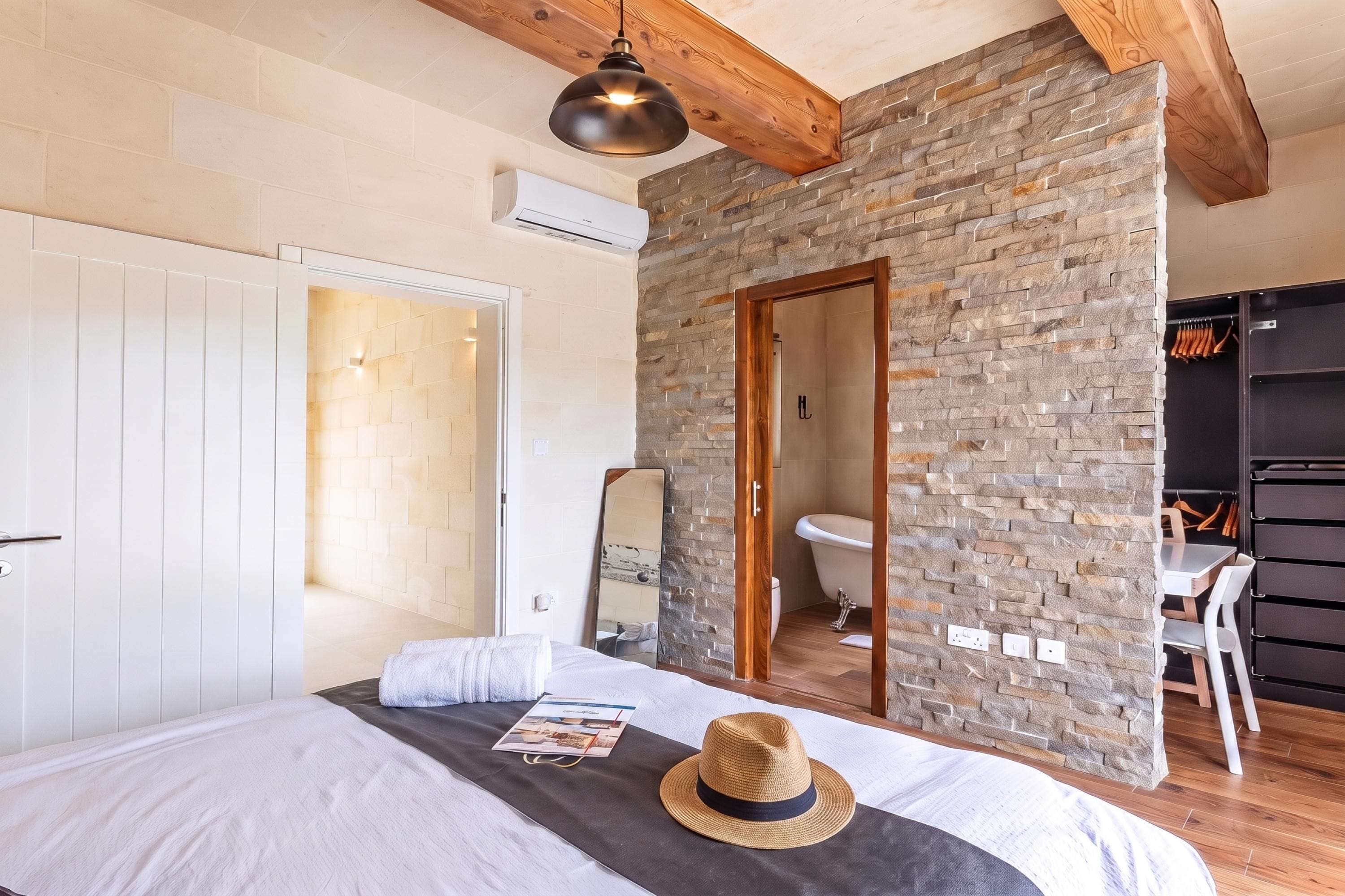 Un dormitorio con una cama grande, techo de madera, pared de piedra, baño con inodoro y lavamanos, y un armario.