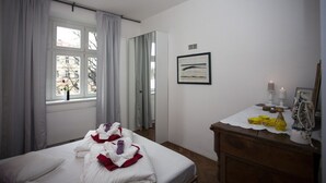 Et hotelværelse med seng, kommode, vindue med gardiner, et billede i ramme på væggen og et lille bord med stearinlys og pynt.